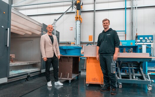 Zwei junge Männer stehen in einer Produktionshalle und lächeln in die Kamera. | © Grote Media/BIS
