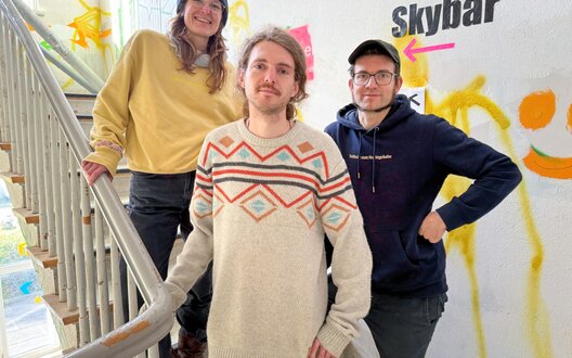 Zwei Männer und eine Frau stehen auf einer Treppe und lächeln. | © WFB/Schack